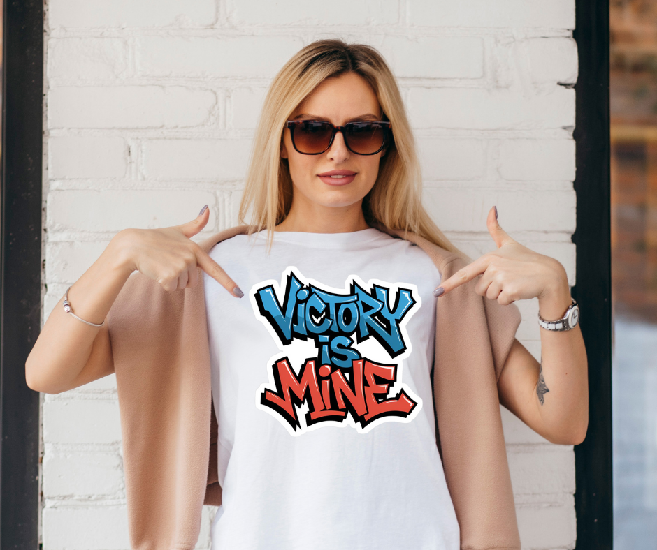 Woman wearing a white t-shirt with 'Victory is Mine' text, pointing at the shirt against a white brick wall.