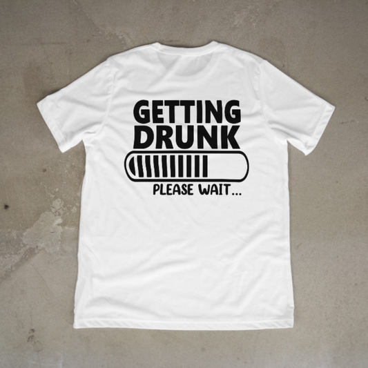 White t-shirt with black text 'GETTING DRUNK PLEASE WAIT...' on a concrete surface