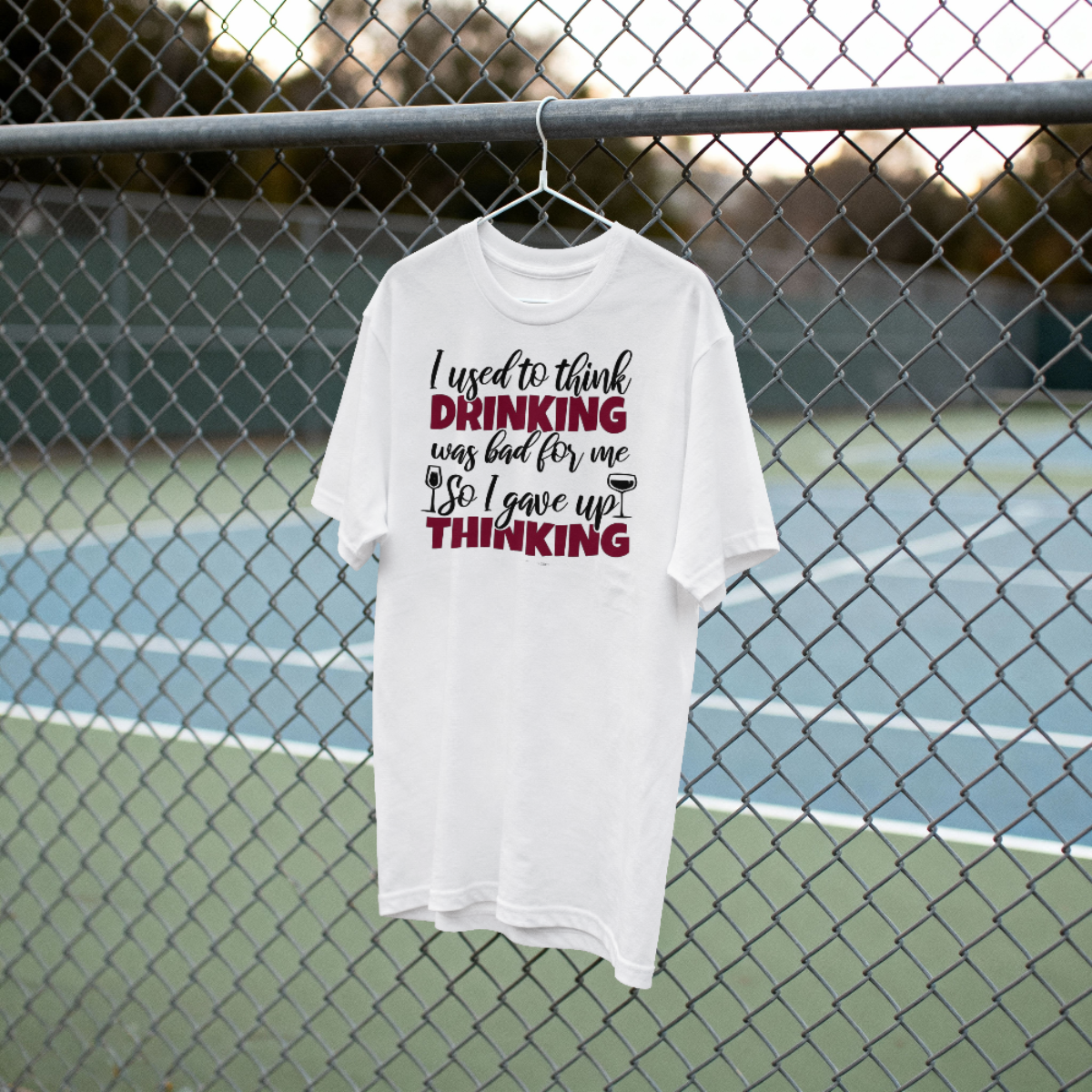 White t-shirt with humorous text design on a hanger against a chain-link fence background.